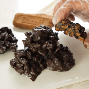 A gloved hand holding a broken chocolate Biscoff cluster to show the biscuit pieces inside.