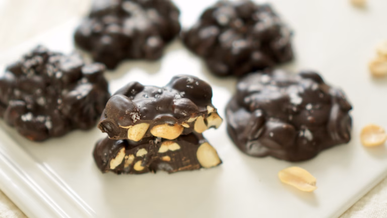 Chocolate peanut clusters on a white plate, with a split cluster showing whole peanuts and a flaky salt topping.