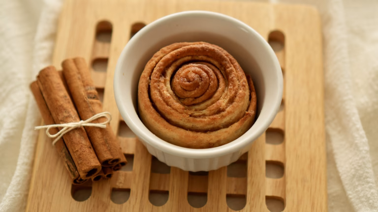 Golden-brown single serve cinnamon roll in a white ramekin beside cinnamon sticks.